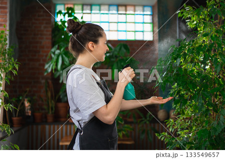 Running of own business. Young woman florist with spray bottle watering fertilizing plants in botanical store. Small business owner working at flower shop smiling surrounded by plants Small business Running of own business. Young woman florist with spray bottle watering fertilizing plants in botanical store. Small business owner working at flower shop smiling surrounded by plants Small business 133549657