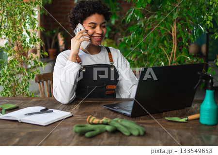 African woman florist gardener working in botanical store using laptop order plants by phone communicating with customer. Female small business owner working at flower shop. Running of own business African woman florist gardener working in botanical store using laptop order plants by phone communicating with customer. Female small business owner working at flower shop. Running of own business 133549658