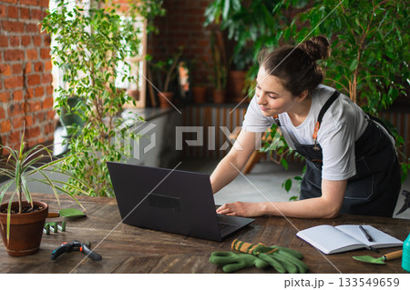 Female florist gardener working in botanical store greenhouse using laptop order plants communicating on Internet with customer. Small business owner working at flower shop. Running of own business Female florist gardener working in botanical store greenhouse using laptop order plants communicating on Internet with customer. Small business owner working at flower shop. Running of own business 133549659