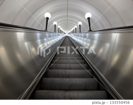 Massive metal escalator leading upward from subway underpass. Numerous steps empty of people. Riding escalator up along path illuminated by lanterns. Tunnel roof round with escalator Massive metal escalator leading upward from subway underpass. Numerous steps empty of people. Riding escalator up along path illuminated by lanterns. Tunnel roof round with escalator 133550456