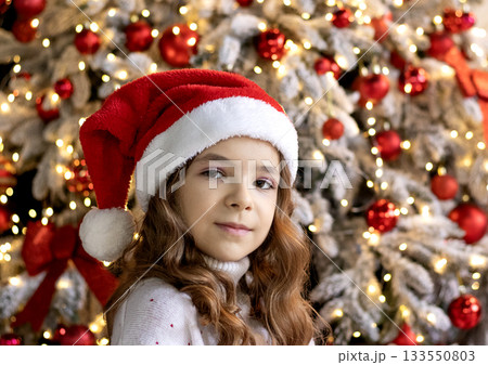 Young girl wearing a Santa hat smiles beside a festive Christmas tree with lights and ornaments 133550803