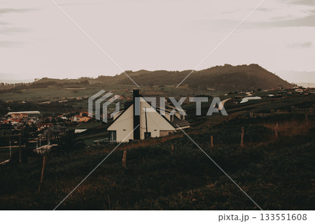 Scenic view of a modern house nestled in a picturesque countryside setting. A contemporary cottage stands out against a backdrop of rolling hills and a charming village, creating a serene landscape. 133551608