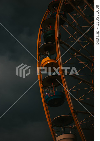 Yellow ferris wheel against a dramatic, cloudy sky. A close-up shot of a observation wheel with colorful cabins against overcast sky. The structure's details are highlighted. Travel and entertainment. 133551609