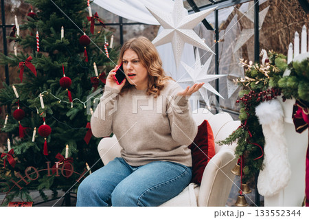 Young angry blonde woman talking on smartphone celebrating Christmas near decorated Christmas tree. 133552344
