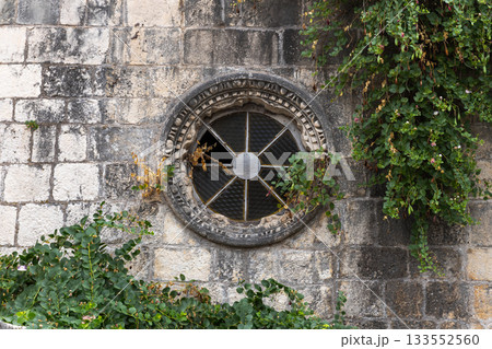 An ancient stone wall features a round, iron-grated window framed by carved stone An ancient stone wall features a round, iron-grated window framed by carved stone 133552560