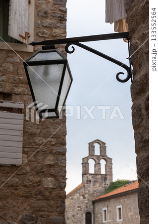 A weathered lantern hangs from a brick wall, framing a distant church tower. Budva 133552564