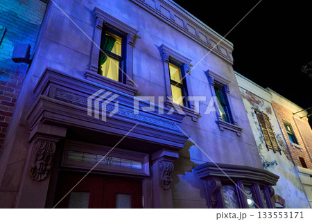Neoclassical stone facade with three glowing windows above a decorated doorway. Blue night lighting contrasts with an adjacent rustic wall and vines. Neoclassical stone facade with three glowing windows above a decorated doorway. Blue night lighting contrasts with an adjacent rustic wall and vines. 133553171