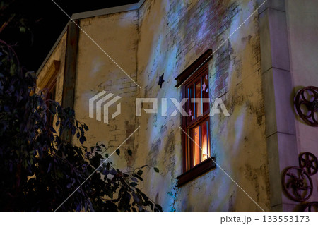 Old plaster facade with a glowing window decorated with star stickers. Night shadows and nearby leaves add moody urban texture. 133553173