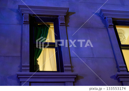 Close-up of a classical window with green drapes and warm interior light. Blue night illumination highlights stone trim and ornate details. Close-up of a classical window with green drapes and warm interior light. Blue night illumination highlights stone trim and ornate details. 133553174