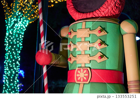 A closeup view shows the green jacket, red belt, and candy cane staff of a giant Elmo nutcracker statue. Holiday lights on nearby trees add colorful bokeh in the night background. A closeup view shows the green jacket, red belt, and candy cane staff of a giant Elmo nutcracker statue. Holiday lights on nearby trees add colorful bokeh in the night background. 133553251