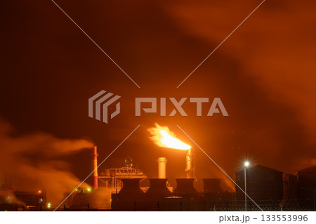 An industrial flare stack burns off excess gas with a large, bright orange flame and thick smoke 133553996
