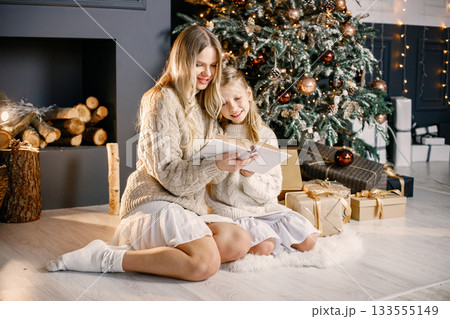 Blonde mother and little daughter sitting near christmas tree at home. Girl with her mother reading a book. Woman and girl wearing white and beige clothes. Blonde mother and little daughter sitting near christmas tree at home. Girl with her mother reading a book. Woman and girl wearing white and beige clothes. 133555149