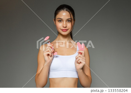 Young woman holding a pink facial roller and a gua sha stone, presenting skincare massage tools during a beauty routine on a neutral studio background. Young woman holding a pink facial roller and a gua sha stone, presenting skincare massage tools during a beauty routine on a neutral studio background. 133558204
