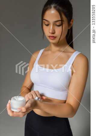 Close-up of a woman applying moisturizing cream to her arm, demonstrating a nourishing skincare step with a jar of body lotion on a neutral background. 133558205