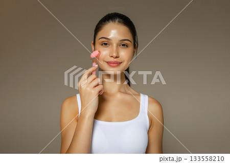 Young woman gently massaging her cheek with a pink facial roller, demonstrating a soothing skincare routine on a neutral studio background. 133558210