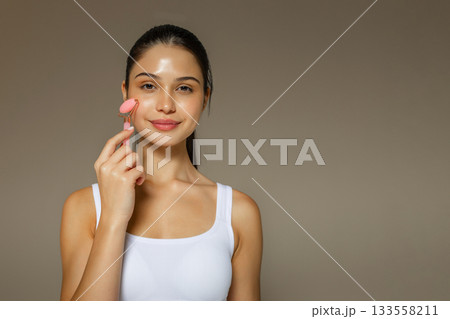 Young woman using a pink facial roller on her cheek, demonstrating a relaxing skincare massage routine on a soft studio background. Young woman using a pink facial roller on her cheek, demonstrating a relaxing skincare massage routine on a soft studio background. 133558211