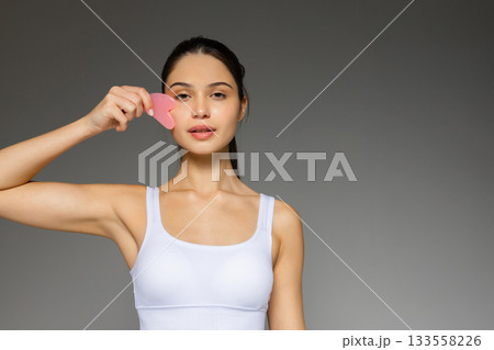 Young woman gently massaging her face with a pink gua sha stone, demonstrating a skincare routine against a neutral background. 133558226