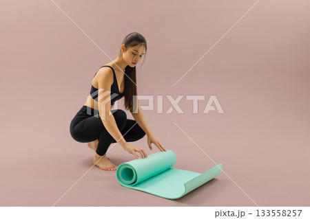 Young woman in black sportswear unrolls a turquoise yoga mat while preparing for a workout on a soft pink background. 133558257