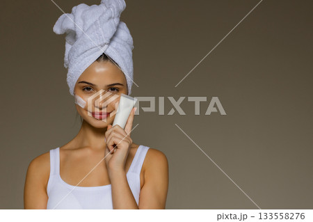 Young woman with a towel on her head applying skincare cream and holding a cosmetic tube, demonstrating a daily beauty routine. 133558276