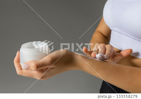 Close-up of a woman applying moisturizing cream to her arm, demonstrating a nourishing skincare step with a jar of body lotion on a neutral background. 133558279