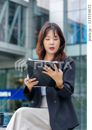 Young Asian woman, business professional, works on street wearing suit, reviewing important corporate documents on clipboard, showing dedicated concentration in busy urban environment. 133559672