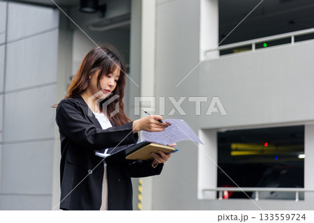 Young Asian business woman in suit busy with work, reviewing important documents and taking notes on city street holding pen and notebook, professional task demonstrating dedicated employee. 133559724