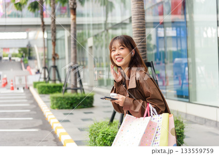 Asian woman enjoys shopping lifestyle, holding colorful fashion bags near modern city mall, smiling, looking away, street with palm trees, people enjoying urban retail during black friday season. 133559759