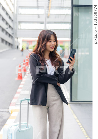 Young Asian business woman in suit stands on street busy with phone smiling confidently ready for work travel with luggage surrounded by modern architecture embodying urban professional life. 133559774