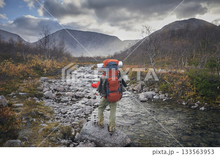 A tourist in a red jacket with a backpack walks by a river in the mountains A tourist in a red jacket with a backpack walks by a river in the mountains 133563953