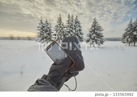 The hand holds the phone. A smartphone is charging from a power bank against the background of a winter landscape with fir trees and a sky with clouds The hand holds the phone. A smartphone is charging from a power bank against the background of a winter landscape with fir trees and a sky with clouds 133563992