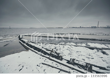 The remains of a wooden ship on a snowy beach river in winter against a gloomy gray sky 133563996