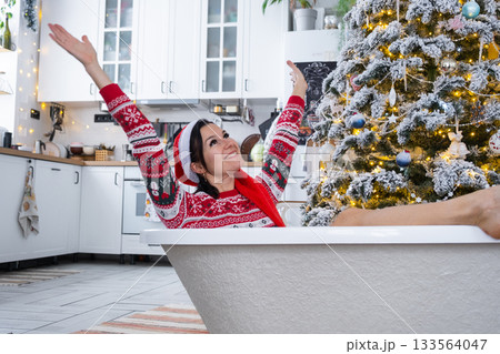 Happy woman in Santa hat is lying resting in New bathtub under Christmas tree as New Year's gift in interior of decorated kitchen. Interior renovation and design, construction and home improvement Happy woman in Santa hat is lying resting in New bathtub under Christmas tree as New Year's gift in interior of decorated kitchen. Interior renovation and design, construction and home improvement 133564047