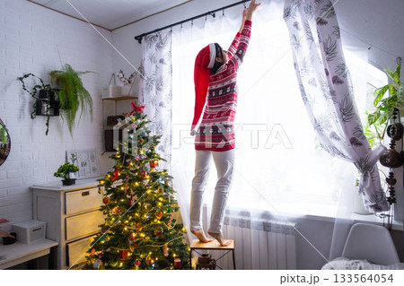 A woman in a Santa hat hangs curtains in the bedroom on the skylights with a Christmas tree and Christmas decorations, preparing for the New Year 133564054