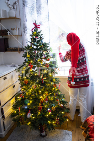 A woman in a Santa hat hangs curtains in the bedroom on the skylights with a Christmas tree and Christmas decorations, preparing for the New Year 133564061