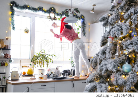 A woman in a Santa hat cleans a kitchen window with Christmas decorations, preparing for the New Year, decorating and tidying up the house A woman in a Santa hat cleans a kitchen window with Christmas decorations, preparing for the New Year, decorating and tidying up the house 133564063