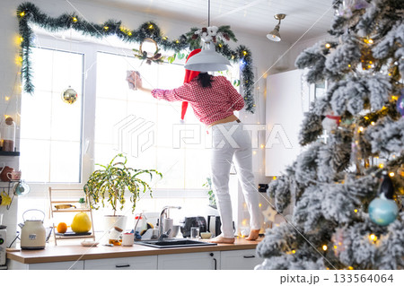A woman in a Santa hat cleans a kitchen window with Christmas decorations, preparing for the New Year, decorating and tidying up the house A woman in a Santa hat cleans a kitchen window with Christmas decorations, preparing for the New Year, decorating and tidying up the house 133564064