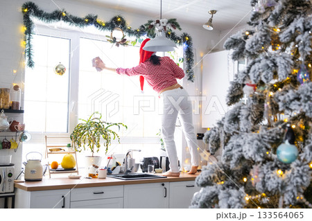 A woman in a Santa hat cleans a kitchen window with Christmas decorations, preparing for the New Year, decorating and tidying up the house A woman in a Santa hat cleans a kitchen window with Christmas decorations, preparing for the New Year, decorating and tidying up the house 133564065