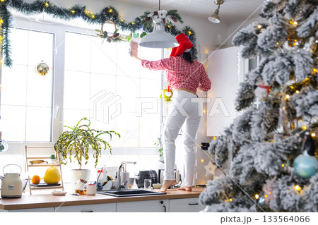 A woman in a Santa hat cleans a kitchen window with Christmas decorations, preparing for the New Year, decorating and tidying up the house A woman in a Santa hat cleans a kitchen window with Christmas decorations, preparing for the New Year, decorating and tidying up the house 133564066