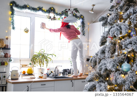 A woman in a Santa hat cleans a kitchen window with Christmas decorations, preparing for the New Year, decorating and tidying up the house A woman in a Santa hat cleans a kitchen window with Christmas decorations, preparing for the New Year, decorating and tidying up the house 133564067