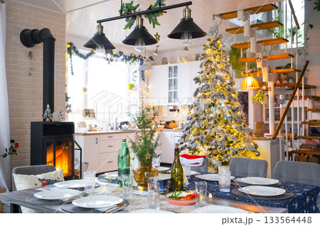 Festive interior of house is decorated for Christmas and New Year in loft style with black stove, fireplace, Christmas tree. Warm studio room with set table, burning wood, cozy and heating of home 133564448