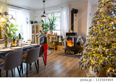 Festive interior of house is decorated for Christmas and New Year in loft style with black stove, fireplace, Christmas tree. Warm studio room with set table, burning wood, cozy and heating of home Festive interior of house is decorated for Christmas and New Year in loft style with black stove, fireplace, Christmas tree. Warm studio room with set table, burning wood, cozy and heating of home 133564464