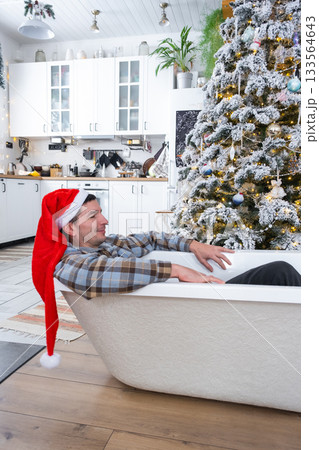 Man in Santa hat is lying resting in New bathtub under Christmas tree as New Year's gift in interior of white decorated kitchen. Interior renovation and design, construction and home improvement Man in Santa hat is lying resting in New bathtub under Christmas tree as New Year's gift in interior of white decorated kitchen. Interior renovation and design, construction and home improvement 133564643
