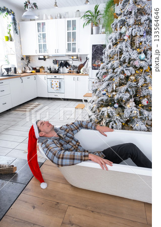 Man in Santa hat is lying resting in New bathtub under Christmas tree as New Year's gift in interior of white decorated kitchen. Interior renovation and design, construction and home improvement Man in Santa hat is lying resting in New bathtub under Christmas tree as New Year's gift in interior of white decorated kitchen. Interior renovation and design, construction and home improvement 133564646