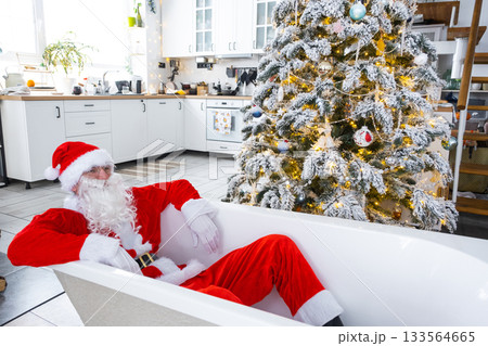 Santa is lying resting in New bathtub under festive Christmas tree as New Year's gift in interior of white modern decorated kitchen. Interior renovation and design, construction and home improvement Santa is lying resting in New bathtub under festive Christmas tree as New Year's gift in interior of white modern decorated kitchen. Interior renovation and design, construction and home improvement 133564665