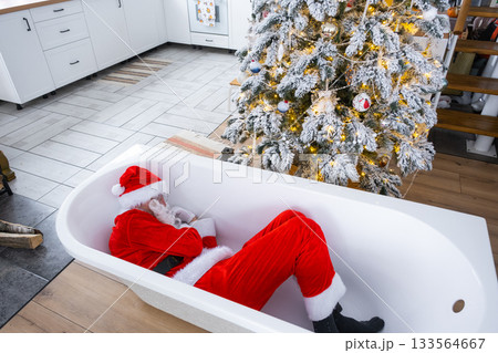 Santa is lying resting in New bathtub under festive Christmas tree as New Year's gift in interior of white modern decorated kitchen. Interior renovation and design, construction and home improvement Santa is lying resting in New bathtub under festive Christmas tree as New Year's gift in interior of white modern decorated kitchen. Interior renovation and design, construction and home improvement 133564667