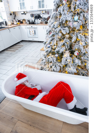 Santa is lying resting in New bathtub under festive Christmas tree as New Year's gift in interior of white modern decorated kitchen. Interior renovation and design, construction and home improvement Santa is lying resting in New bathtub under festive Christmas tree as New Year's gift in interior of white modern decorated kitchen. Interior renovation and design, construction and home improvement 133564669