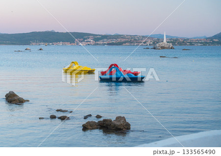 Two colorful pedal boats float on a calm blue sea during early evening light. Soft tones and gentle ripples create a relaxing coastal atmosphere with distant shoreline and rocks in view. 133565490