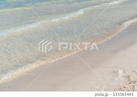 A small wave gently reaches the sandy beach, showcasing the clarity of pale turquoise sea water. The simple natural composition reflects calmness and a serene coastal moment. A small wave gently reaches the sandy beach, showcasing the clarity of pale turquoise sea water. The simple natural composition reflects calmness and a serene coastal moment. 133565491