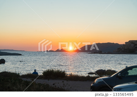 A vivid sunset descends over the sea, illuminating the coastline with warm orange tones. A parked car on the shore adds a touch of everyday life to the tranquil seaside landscape 133565495