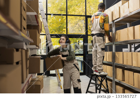 Small depot employee moves cardboard boxes with music on headset, wearing coveralls and preparing parcels for shipment. Arranging goods on shelving units for easy e-shop distribution. 133565727